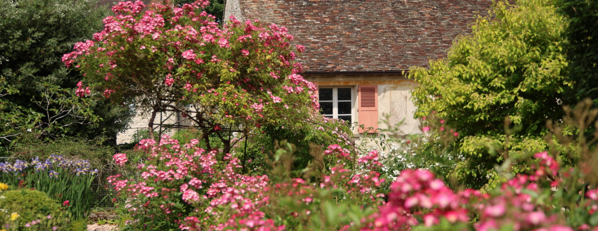 Photographie d'un jardin représentant une maison en pierre ancienne avec des fleurs au premier plan et une allée gravillonnée