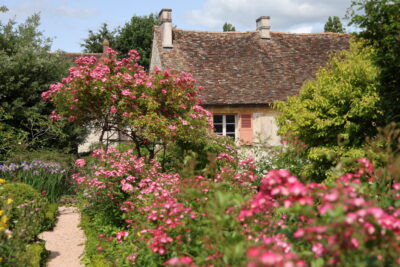 Photographie d'un jardin représentant une maison en pierre ancienne avec des fleurs au premier plan et une allée gravillonnée - Agrandir l'image 7 sur 9, fenêtre modale